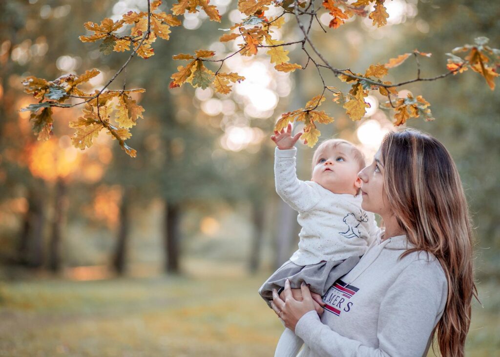 Une femme tient son bébé en l’air dans un parc, capturant le moment pour expliquer comment devenir photographe.