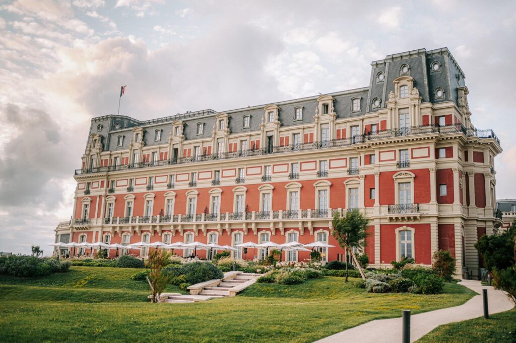 Un grand bâtiment rouge sur un espace vert herbeux : l'Hôtel du Palais à Biarritz.