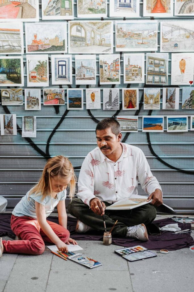 Un homme et une petite fille assis par terre devant un mur de peintures pendant leurs vacances à Porto.