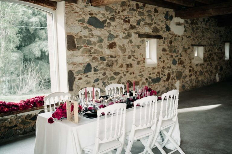 Une table avec des chaises blanches et des fleurs magenta devant un mur de pierre, source d'inspiration pour un mariage.