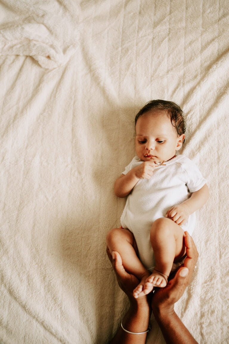 Un bébé est assis sur un lit, les mains sur les genoux.