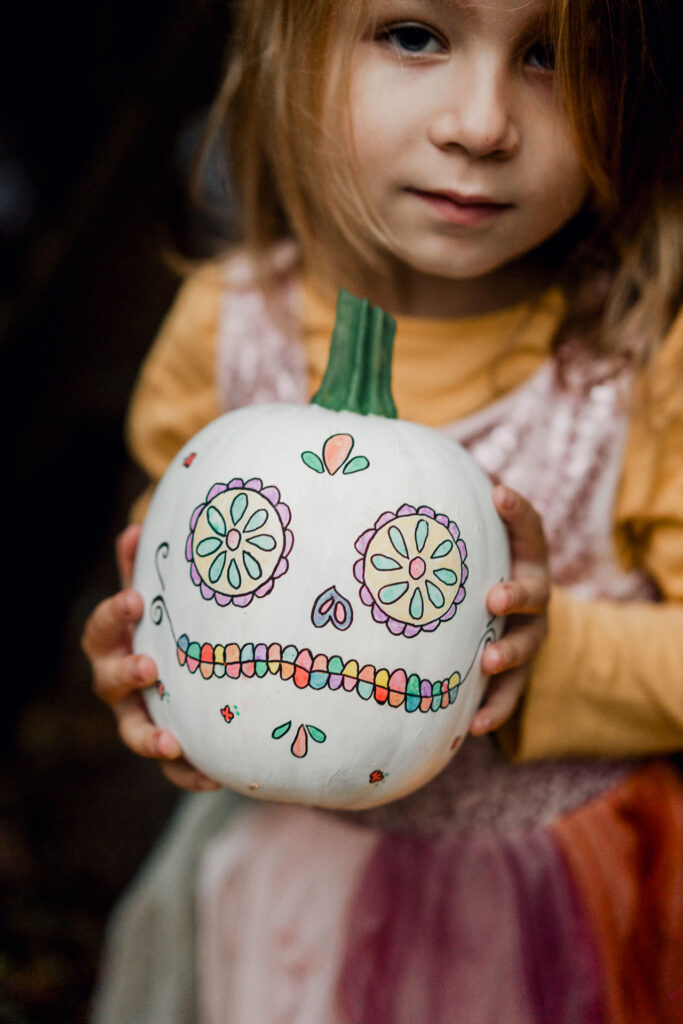 une petite citrouille blanche est dessiné facçon masque mexicain et est tenue dans des mains d'enfants.