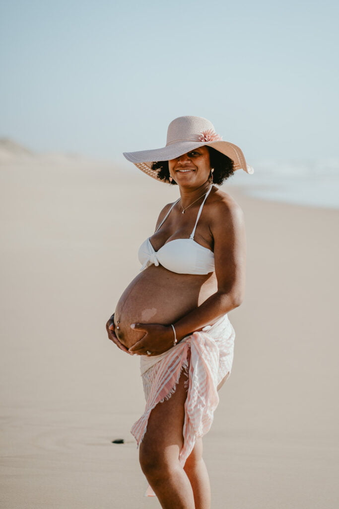 Une femme enceinte de couleur de peau noire sur la plage de Capbreton, avec un chapeau.