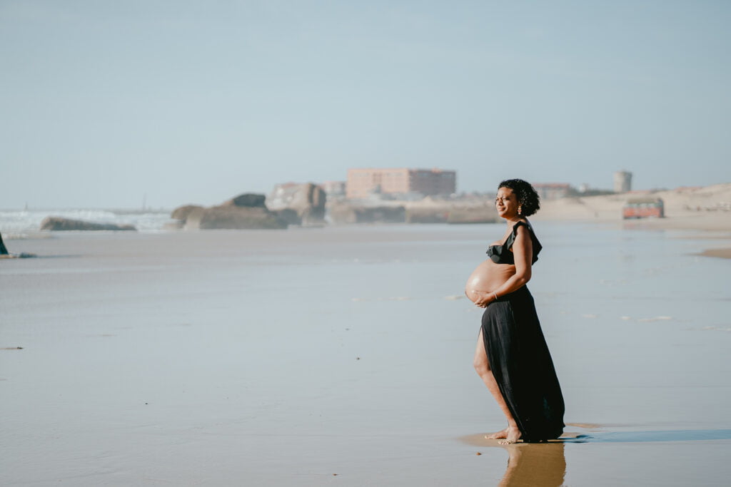 Une femme enceinte vêtue d’une robe noire debout sur la plage.