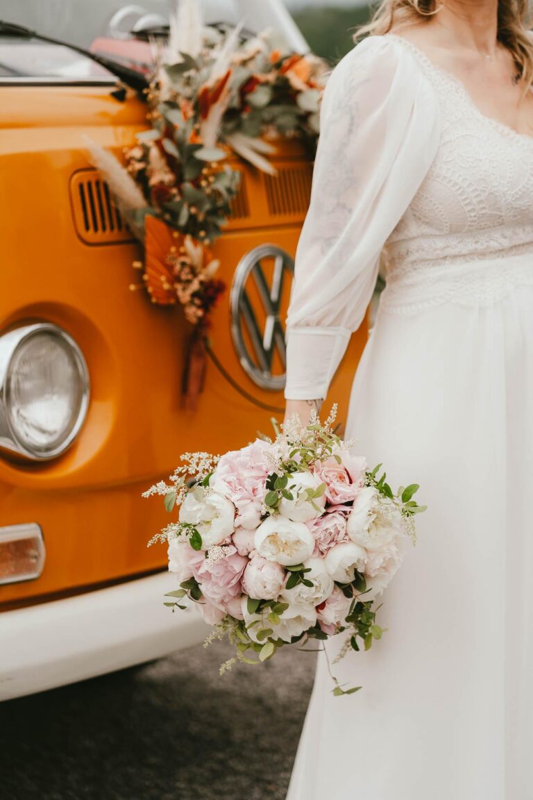 Une mariée en robe blanche debout à côté d'un bus VW orange décoré par un pau de mariage fleuriste.