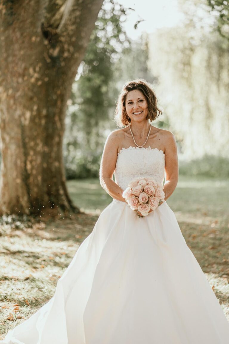 Une mariée en robe de mariée blanche, décorée par un fleuriste mariage landais, debout sous un arbre.