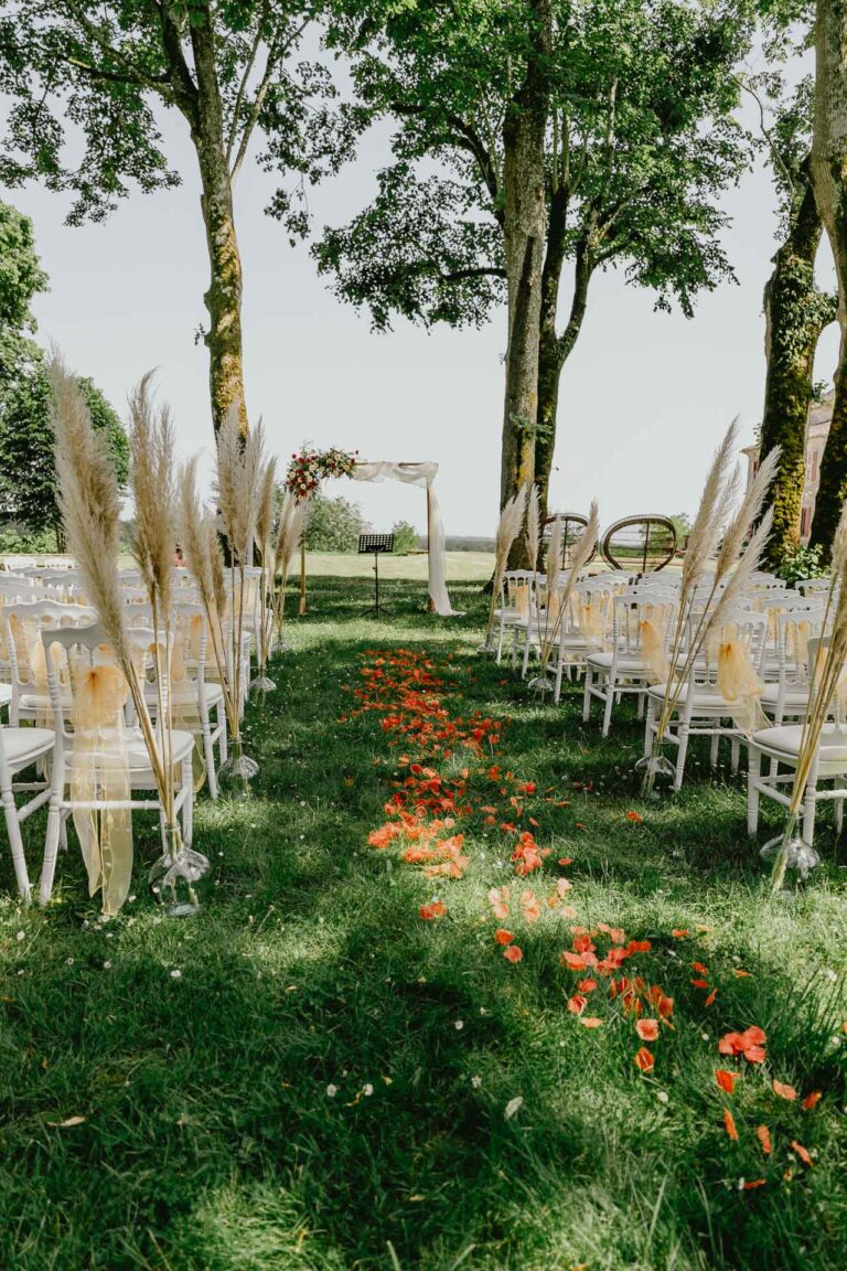 Une cérémonie en plein air aménagée avec des chaises blanches et des plumes oranges, incluant location matériel mariage Landes.