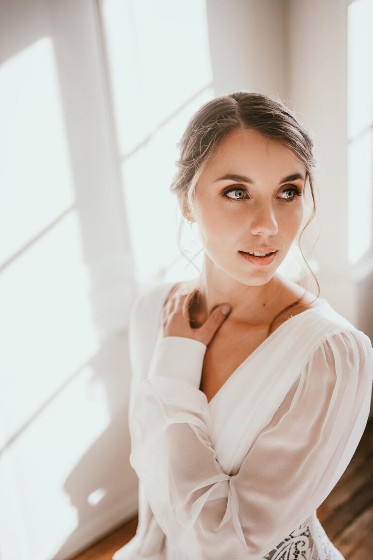 Une belle mariée en robe blanche, savamment préparée par une maquilleuse mariage landaise, posant devant une fenêtre.