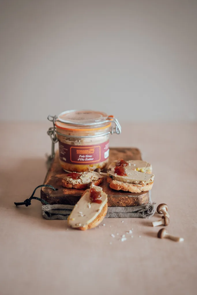 Un pot de foie gras est posé sur une planche en bois avec deux tartines de pain, pour un shooting culinaire.