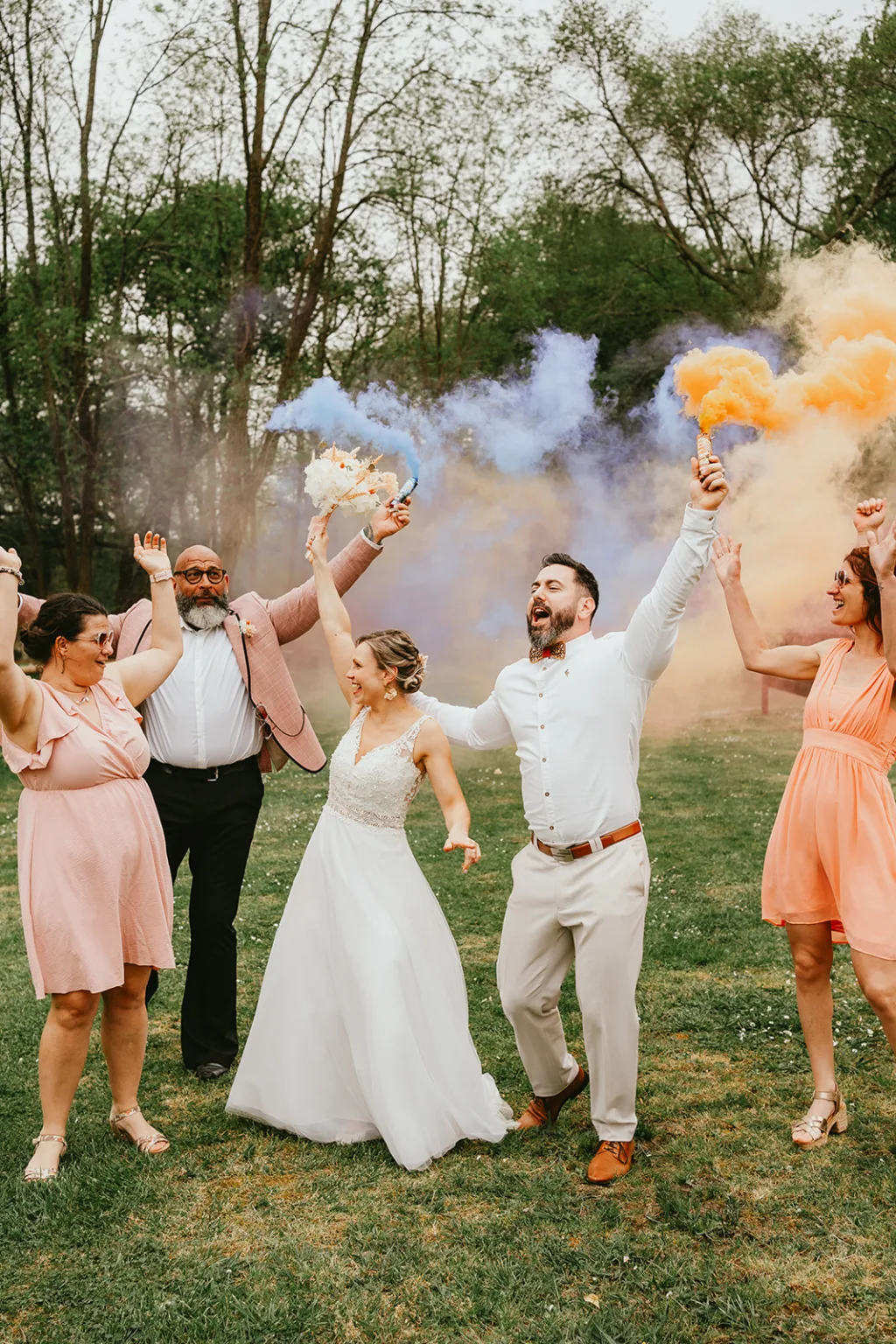 Un groupe de personnes, dont des mariés, célèbrent à l’extérieur en tenant des bombes fumigènes émettant une fumée magenta colorée. L’ambiance est joyeuse et festive, parfaite pour des moments de mariage inspirants.