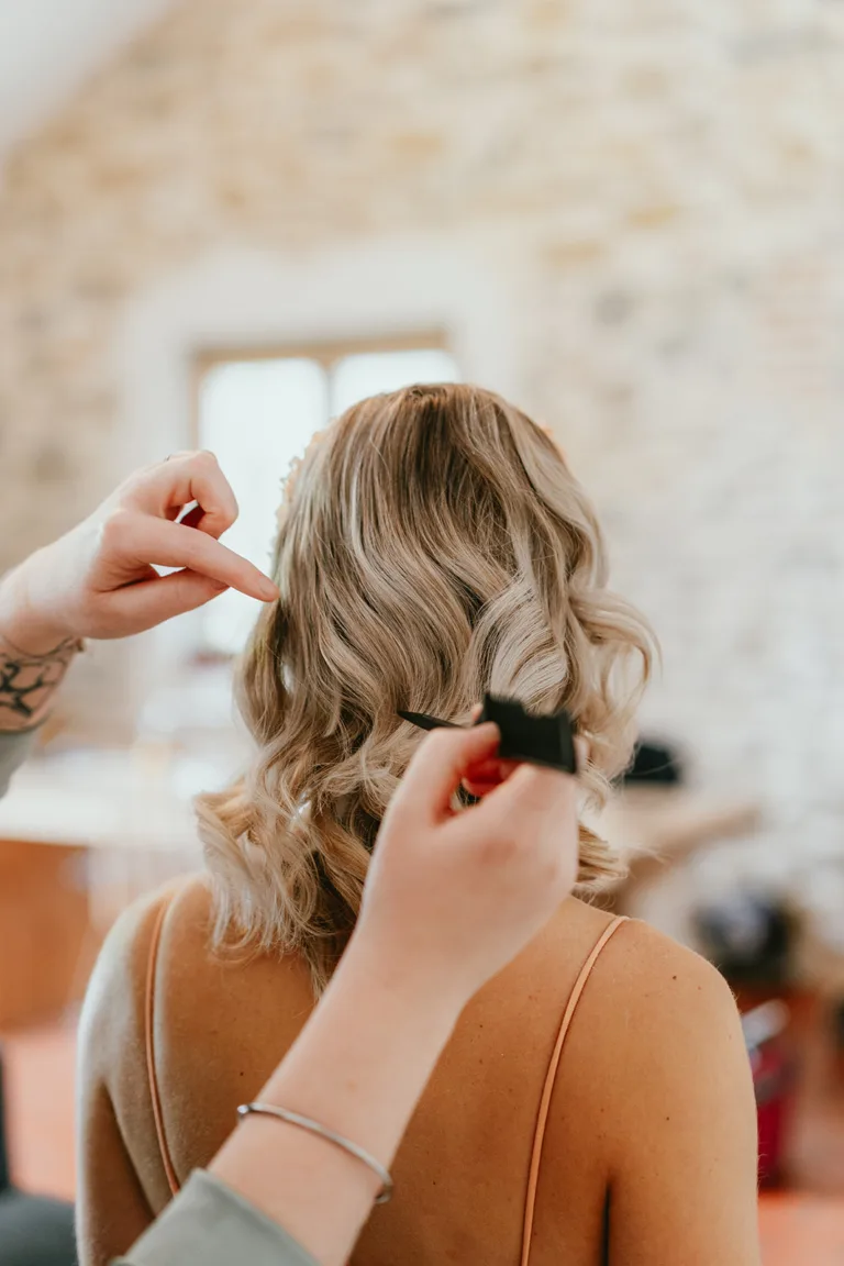 Un coiffeur ajuste les cheveux blonds et ondulés d'une femme avec un fer à friser dans une pièce aux murs en pierre, s'inspirant des styles de mariage élégants, tandis que les accents magenta ajoutent une touche de couleur au cadre sophistiqué.