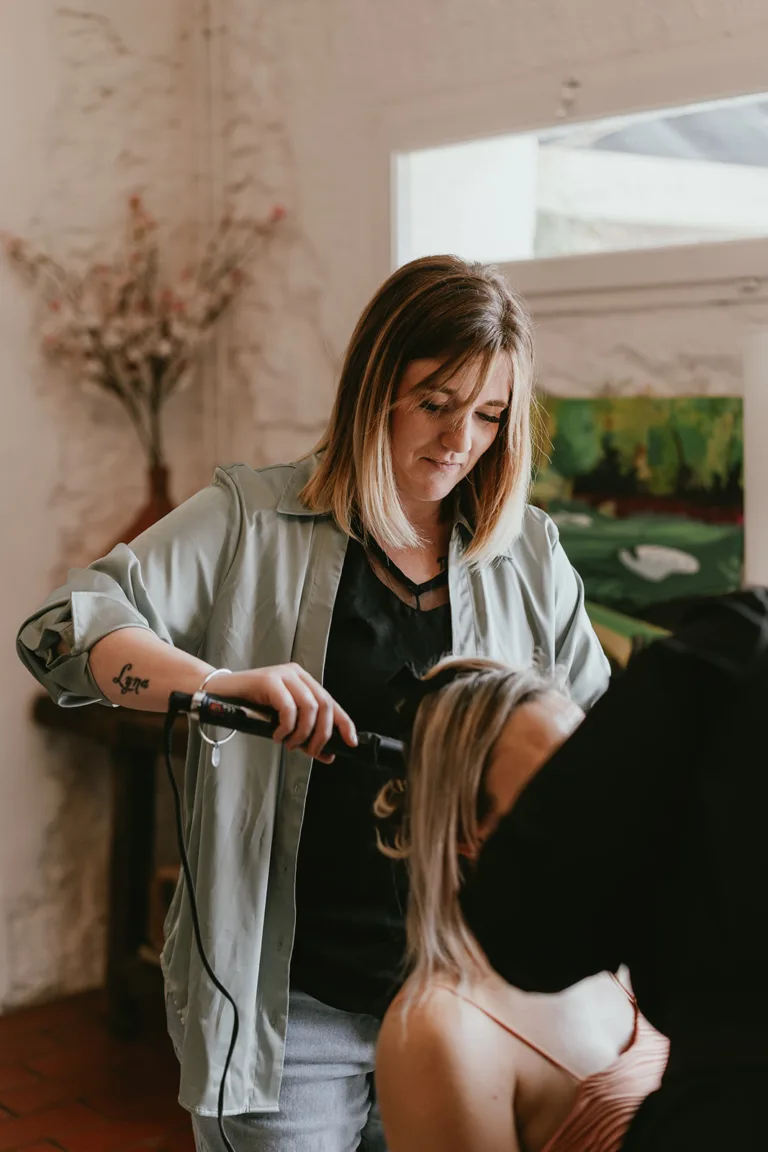 Une coiffeuse aux cheveux bruns et tatouée sur l'avant-bras boucle les cheveux d'une cliente dans un salon bien éclairé, s'inspirant d'un magnifique tableau orné de fleurs magenta en arrière-plan.