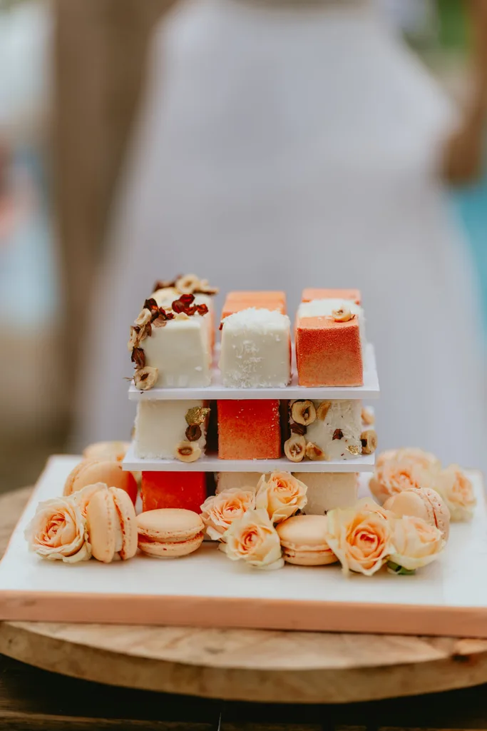 Présentoir à desserts à trois niveaux avec gâteaux carrés et macarons, orné de roses orange pâle pour un mariage d'inspiration, posé sur une table en bois.