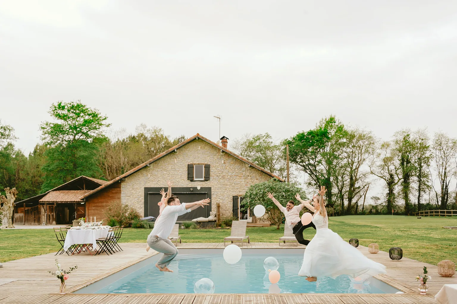 Deux personnes sautent dans une piscine avec des ballons magenta ; une table est dressée pour dîner à proximité, et une maison en pierre et des arbres verts en arrière-plan inspirent une célébration de mariage parfaite.