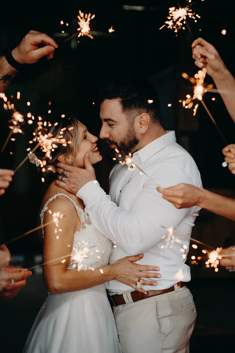 Un couple en tenue de mariage s'embrasse et se sourit, entouré de personnes tenant des cierges magiques, créant un moment de pure inspiration mariage contre l'éclat enchanteur des teintes magenta.