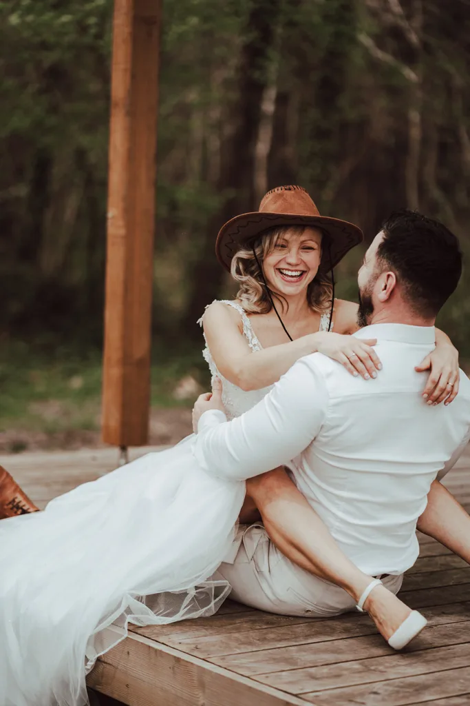 Une femme vêtue d'une robe blanche et d'un chapeau de cowboy est assise sur les genoux d'un homme en tenue blanche sur une terrasse en bois. Tous deux sourient, se tiennent la main et se regardent dans les yeux comme s'ils étaient capturés sur une photo de séance éthérée.