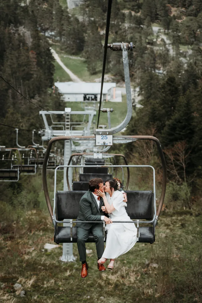Un couple en costume vert et robe blanche est assis sur un téléski et partage un baiser. L’arrière-plan présente une forêt dense et un terrain vallonné, créant la séance parfaite pour un moment magique.