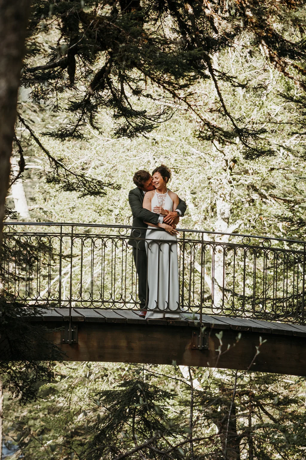Un couple en tenue de soirée se tient sur un pont métallique entouré d'arbres, l'homme serrant la femme par derrière, capturant un moment photo de famille parfait.