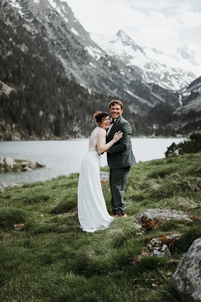 Des mariés en tenue de mariage formelle se tiennent sur une zone herbeuse au bord d'un lac avec des montagnes et de la neige en arrière-plan, souriant et s'enlaçant comme s'ils capturaient une séance extraordinaire.