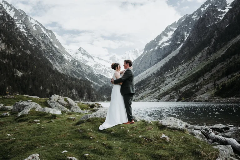 Un couple en tenue de mariage s'embrasse sur une zone herbeuse au bord d'un lac, entouré de montagnes enneigées sous un ciel partiellement nuageux, comme s'il capturait une photo de famille sereine.