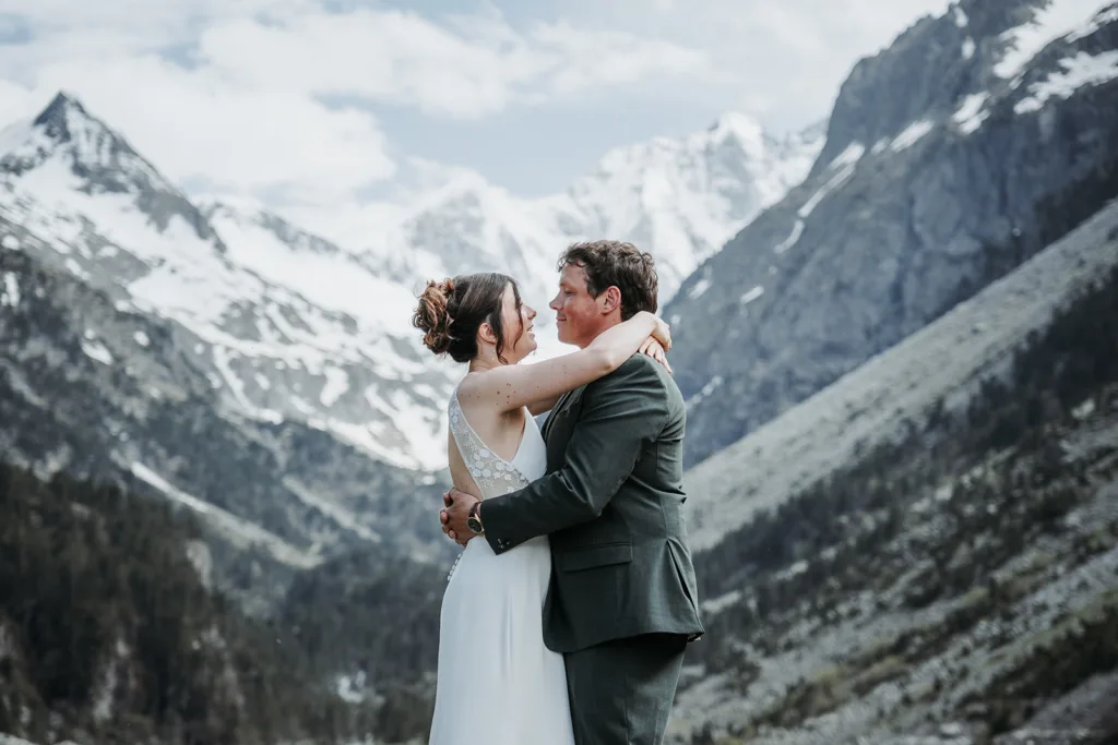 Un couple en tenue de mariage s'embrasse devant un paysage montagneux pittoresque avec des sommets enneigés et un ciel nuageux, créant une photo intemporelle rappelant les séances de famille chéries.