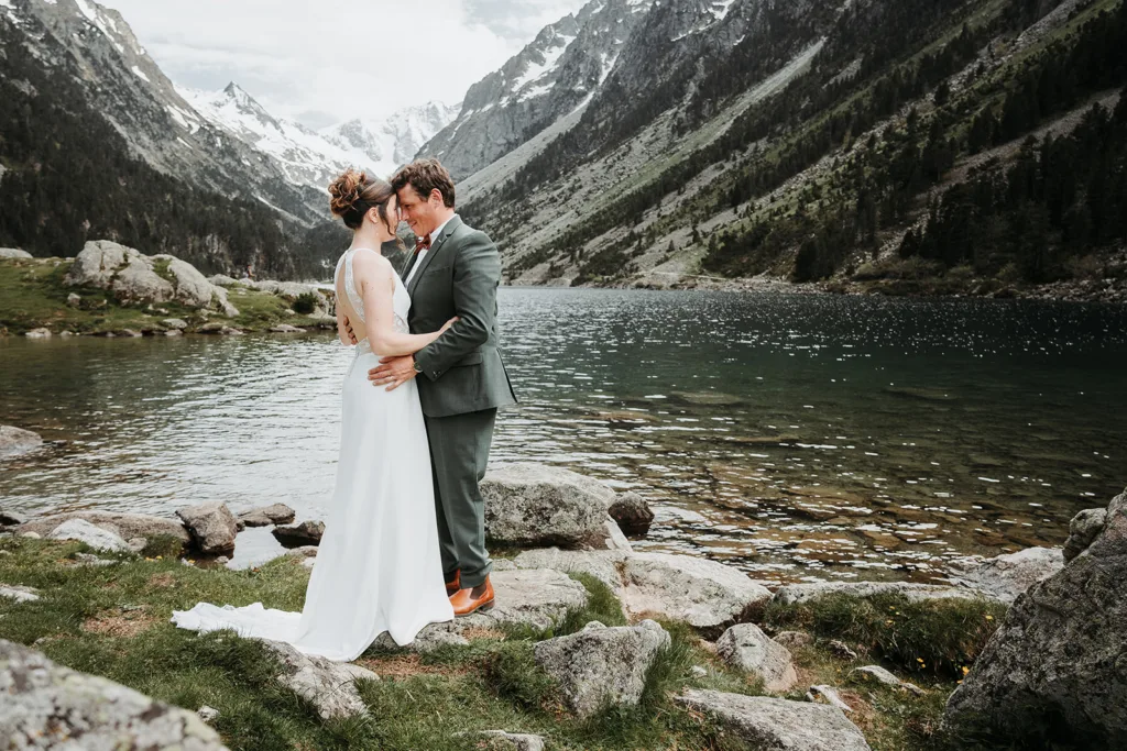 Un couple en tenue de mariage s'embrasse près d'un lac de montagne tranquille, avec des sommets enneigés et une verdure luxuriante en arrière-plan, créant une séance de romance intemporelle.