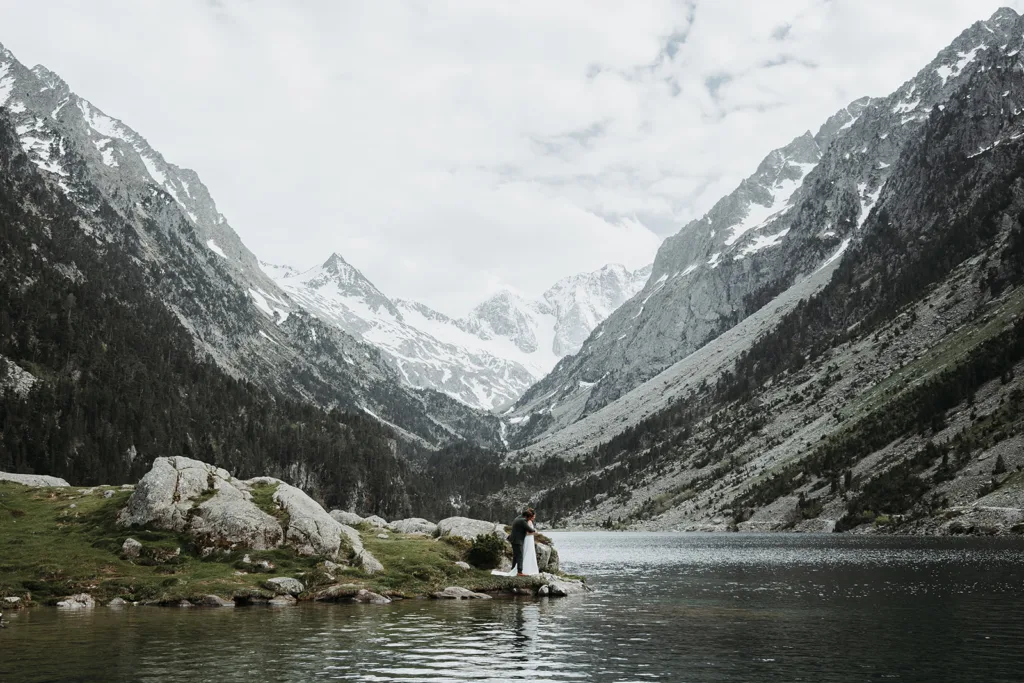 Une personne se tient au bord d’un lac de montagne calme, entouré de falaises rocheuses et de sommets enneigés sous un ciel nuageux, rappelant une sereine photo de famille.