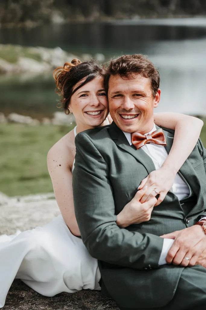 Un couple en tenue de mariage sourit et s'embrasse alors qu'il est assis à l'extérieur près d'un lac serein, créant ainsi une photo de famille chérie.