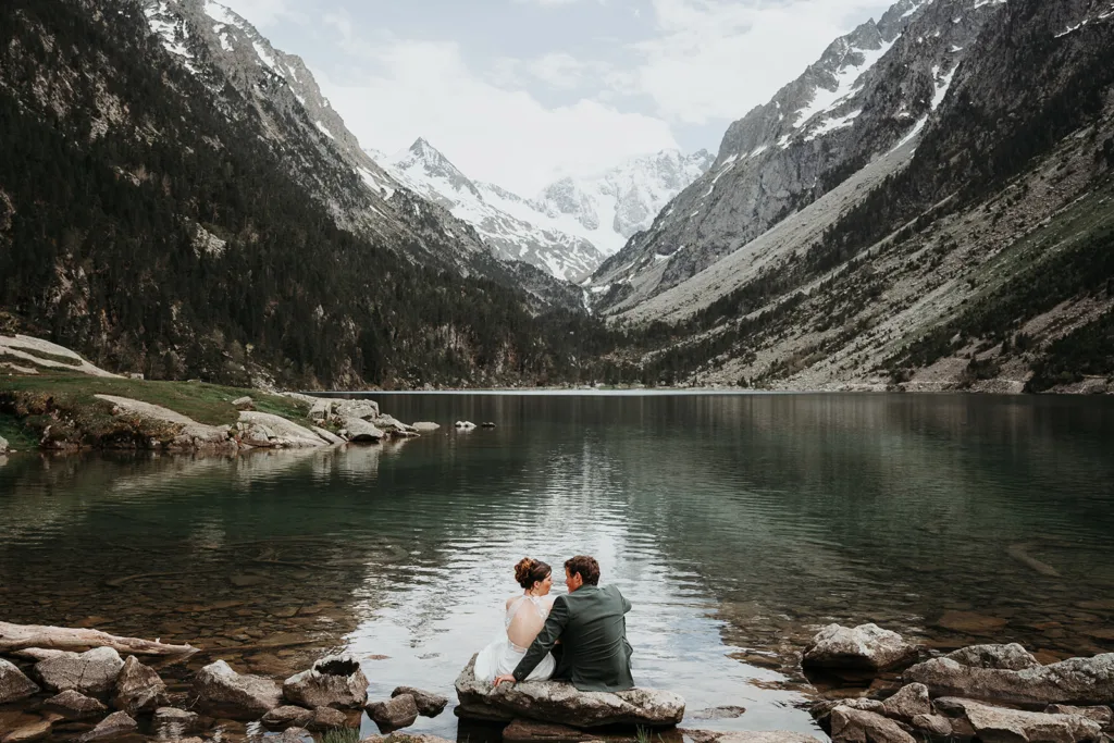 Un couple est assis sur un rocher au bord d'un lac de montagne serein, entouré de sommets enneigés et de verdure luxuriante, capturant l'instant comme s'il s'agissait d'une belle séance photo.