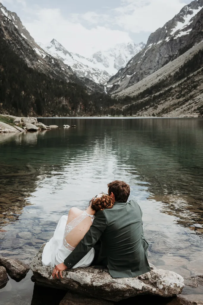Un couple en tenue formelle est assis sur un rocher au bord d’un lac, entouré de montagnes et de sommets partiellement enneigés. La femme pose sa tête sur l'épaule de l'homme alors qu'ils font face au paysage serein, capturant une séance intime rappelant une photo de famille intemporelle.