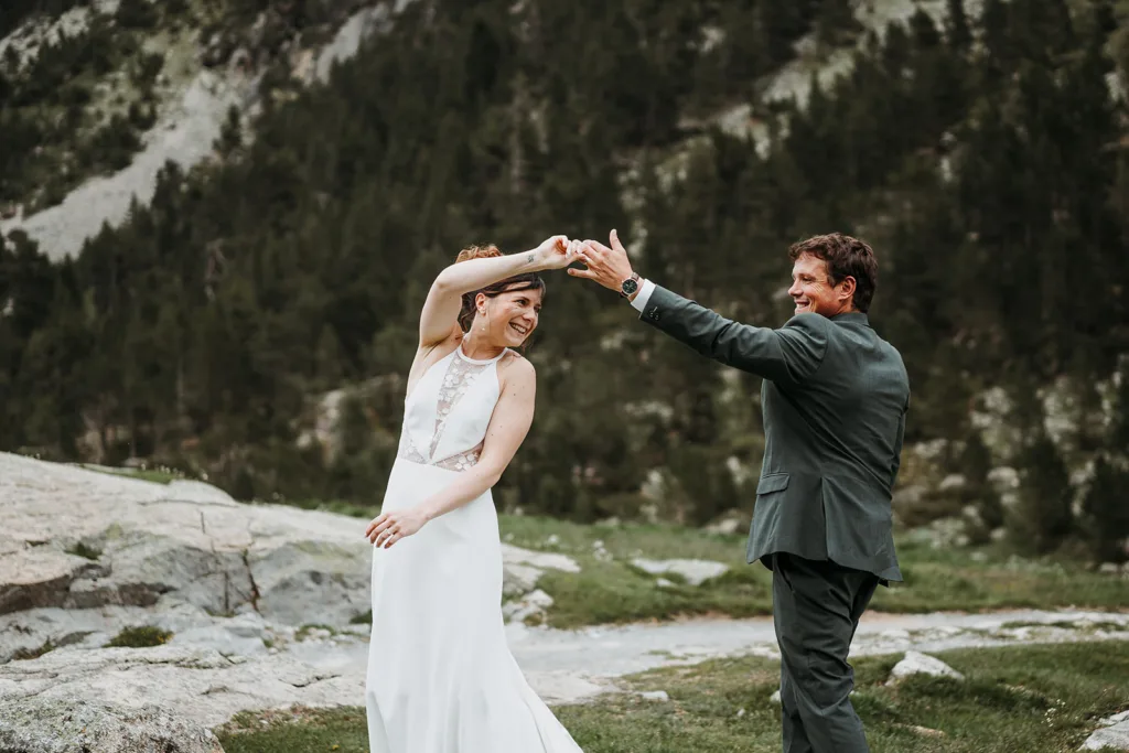 Un couple, vêtu d'une tenue de mariage, danse joyeusement à l'extérieur sur une zone herbeuse avec des arbres et un terrain rocheux en arrière-plan, créant un moment photo de famille parfait.