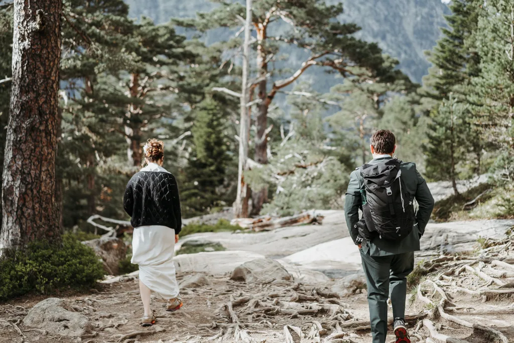 Deux personnes, l'une vêtue d'une robe blanche et l'autre portant une veste noire avec un sac à dos, font une randonnée sur un sentier forestier entouré d'arbres et de terrains rocheux, comme capturées lors d'une séance impromptue au milieu de la beauté brute de la nature.