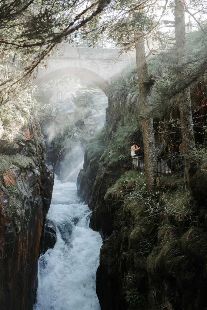 Une rivière rugissante coule à travers une gorge rocheuse entourée d'arbres moussus, avec un pont au-dessus. Deux personnes se tiennent sur une petite plate-forme près de la rivière, admirant la vue et prenant une photo de séance pour se souvenir de ce moment serein.