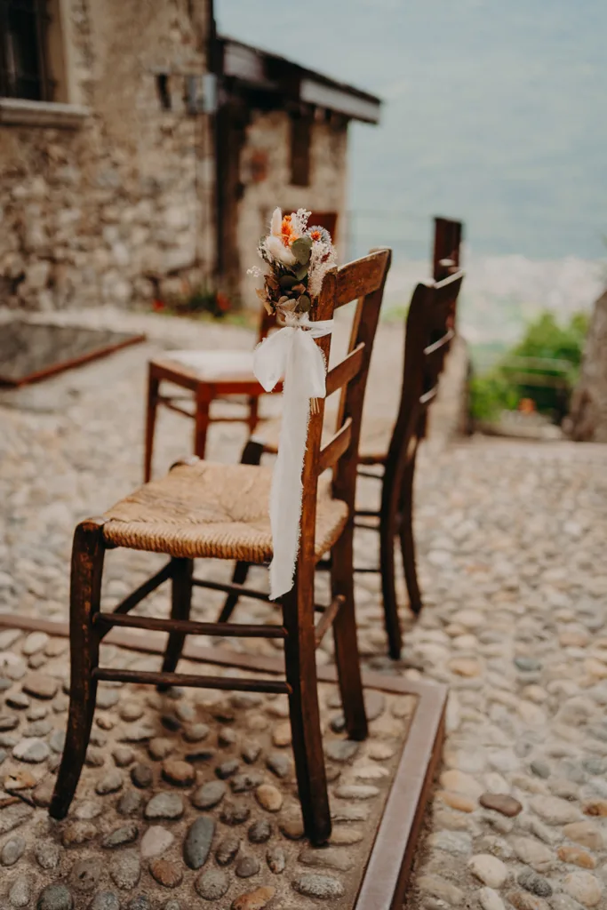 Deux chaises en bois aux décorations florales et aux rubans blancs sont placées sur une allée en pierre à l'extérieur d'un bâtiment rustique, parfait pour un mariage en Italie.