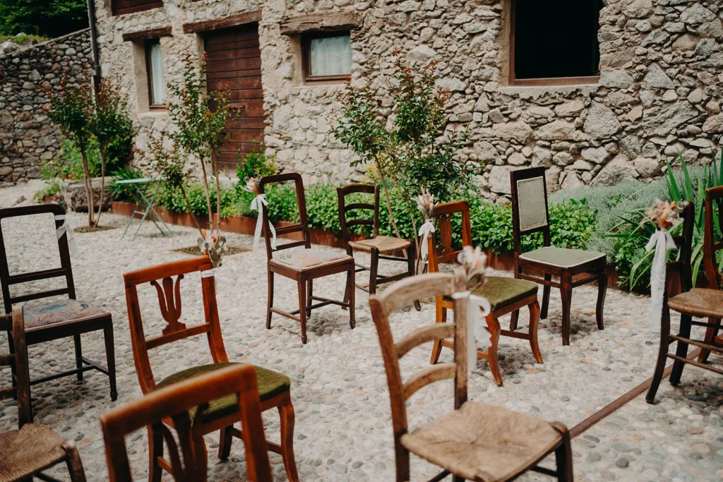 Une configuration de mariage en plein air avec des chaises en bois assorties disposées sur une surface pavée devant un bâtiment en pierre, chaque chaise ornée de petites décorations florales, capturant l'essence d'un mariage en Italie.