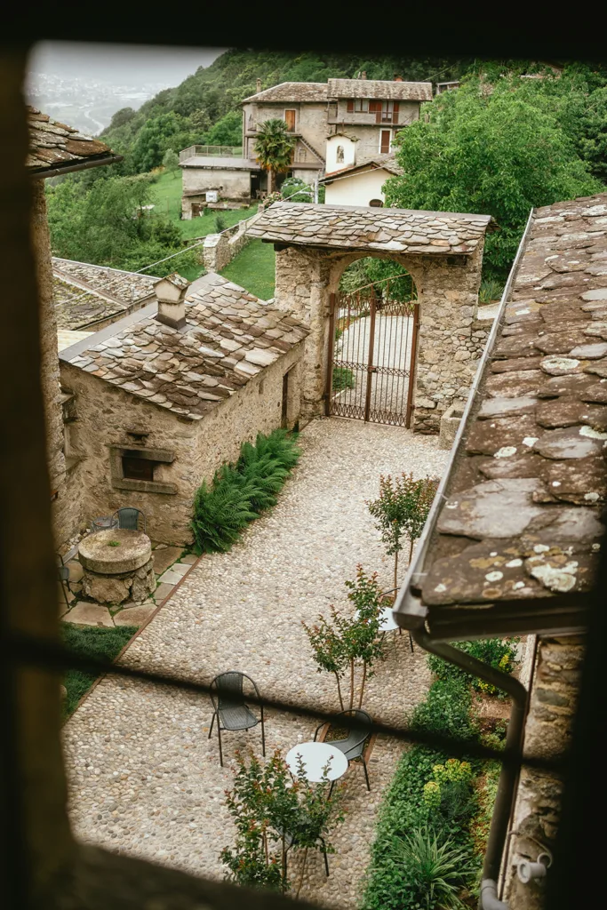 Vue depuis une fenêtre d'une cour en pierre avec tables et chaises, entourée de bâtiments et de verdure, avec un portail qui s'ouvre sur un paysage pittoresque à flanc de colline : le cadre idéal pour un mariage en Italie.