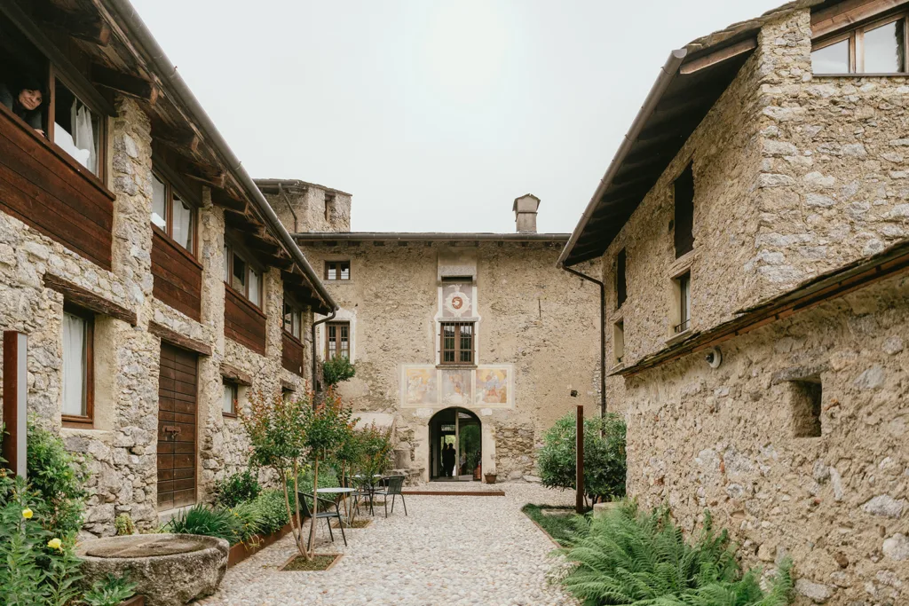 Une cour en pierre avec terrasse est entourée de bâtiments rustiques avec des accents en bois et de petites fenêtres, parfaits pour un charmant mariage en Italie. Une personne regarde depuis une fenêtre supérieure à gauche.