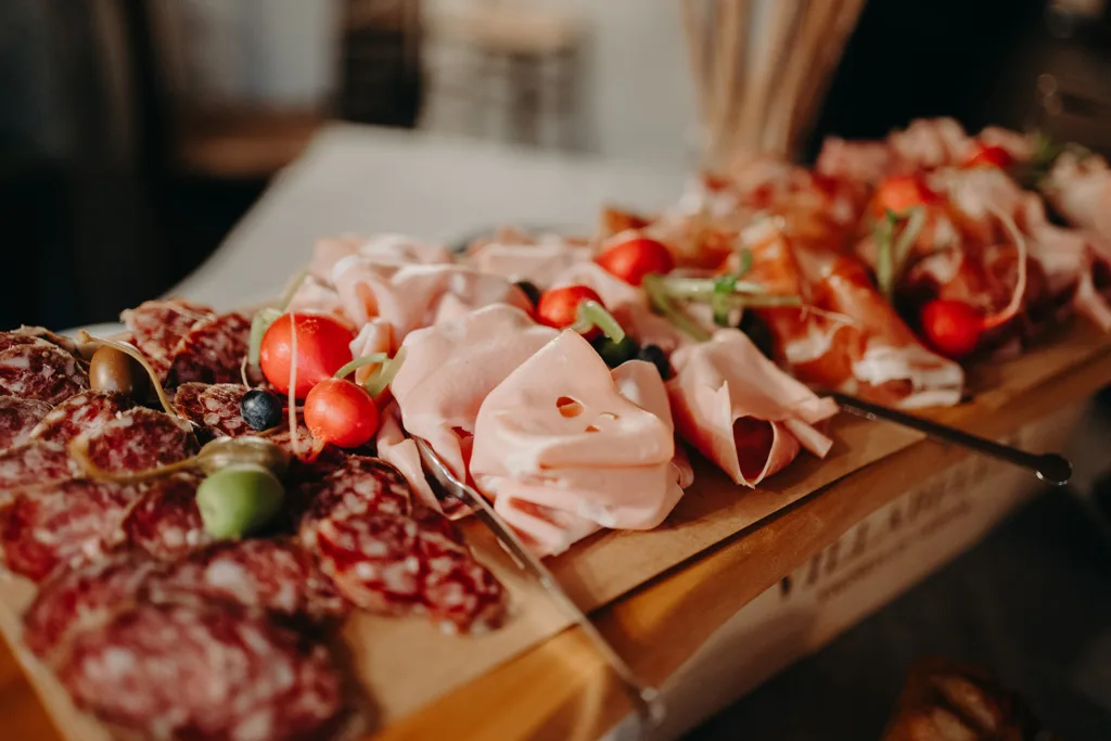 Une planche en bois présentant un assortiment de tranches de viande, dont du salami et du jambon, garnies de tomates cerises, d'olives et d'herbes, une touche parfaite pour un mariage en Italie.