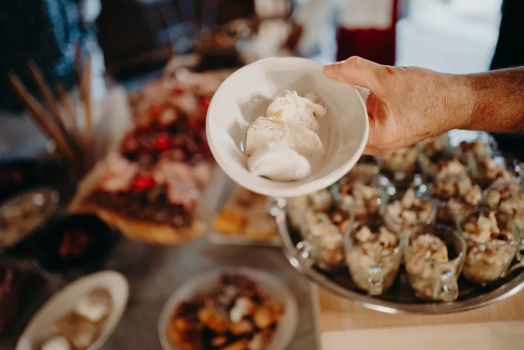 Une personne tient un bol contenant quelques boules de glace. En arrière-plan, une variété de plats, rappelant un délicieux mariage en Italie, sont présentés sur une table.