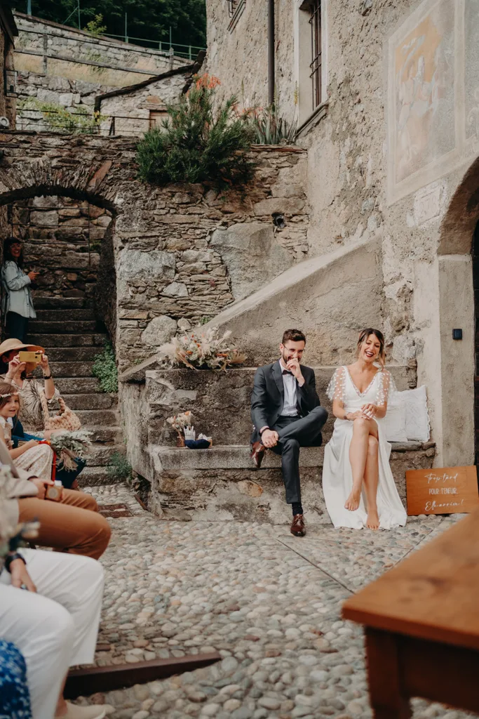 Un couple est assis sur un banc dans une cour extérieure rustique en pierre, entouré d’invités. La femme porte une robe blanche et l'homme est en costume. C'est une scène pittoresque de mariage en Italie, avec des invités assis et regardant sous différents angles.