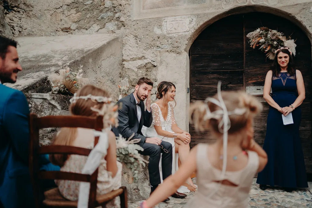 Des mariés sont assis devant un bâtiment rustique lors d’une cérémonie de mariage. Une femme tient des papiers et deux enfants aux cheveux tressés et aux rubans sont au premier plan, capturant le charme d'un mariage en Italie.