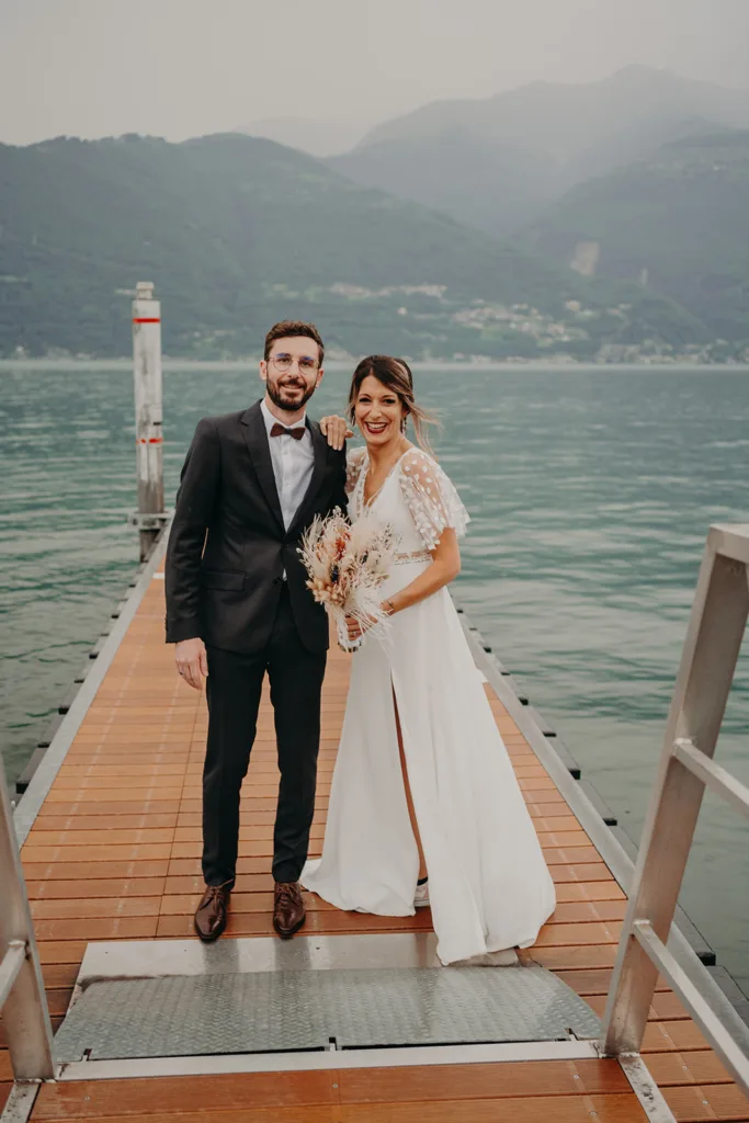 Un couple se tient sur un quai en bois devant un plan d’eau serein avec les majestueuses Pyrénées en arrière-plan. La femme, radieuse dans sa robe de mariée et tenant un bouquet, se tient à côté de l’homme élégamment vêtu d’un costume, capturant une photo de séance intemporelle.