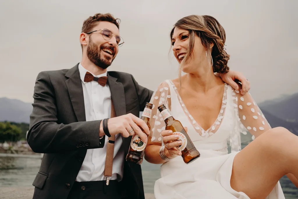 Un homme en costume et une femme en robe blanche partagent un rire tout en tintant des bouteilles de bière en plein air, comme un exemple de joie dans le magnifique décor des Pyrénées.