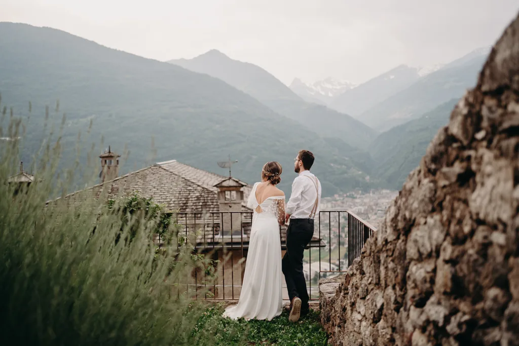 Un couple en tenue de mariage se tient sur une terrasse surplombant les Pyrénées, avec les toits du village au premier plan, un cadre parfait pour ce moment photo de séance.