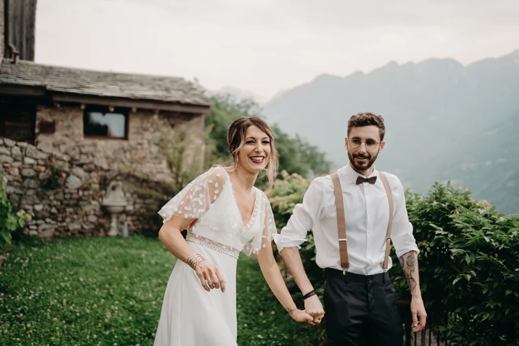 Un couple, habillé en tenue de mariage, se tient la main et sourit en plein air avec un bâtiment rustique en pierre et les majestueuses Pyrénées en arrière-plan.