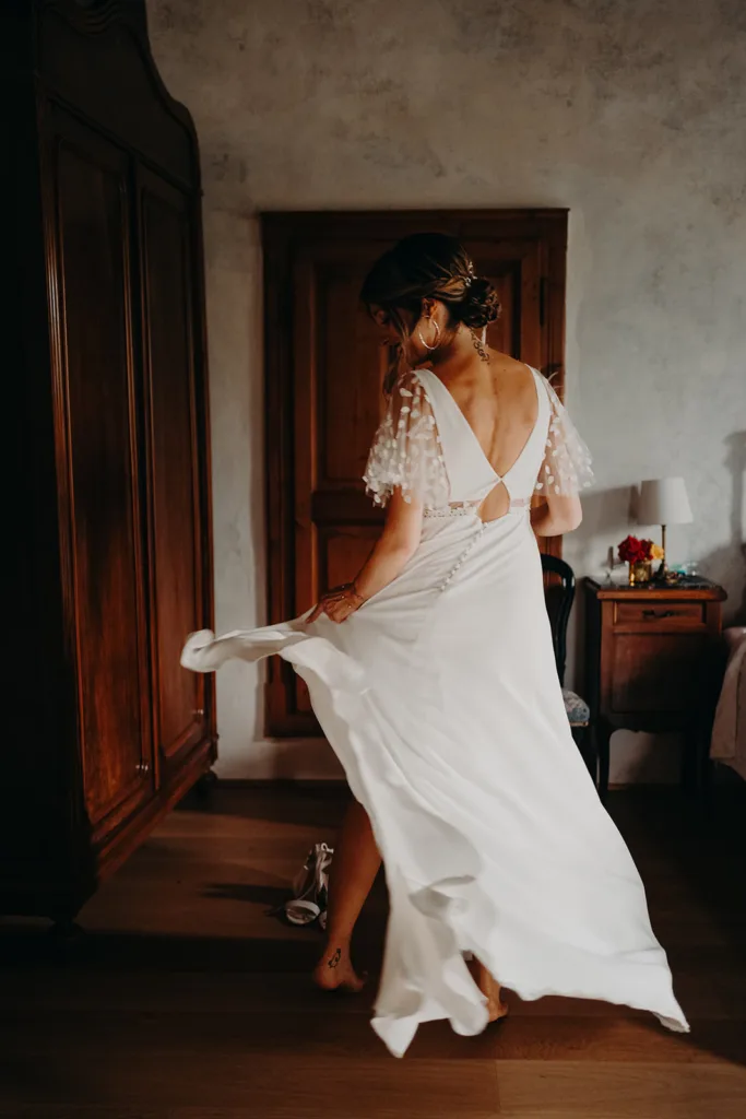 Une femme vêtue d’une robe blanche avec un dos décolleté virevolte gracieusement dans une pièce faiblement éclairée, ressemblant à une scène enchanteresse d’une vieille photo de séance, entourée de meubles en bois et de murs gris clair.