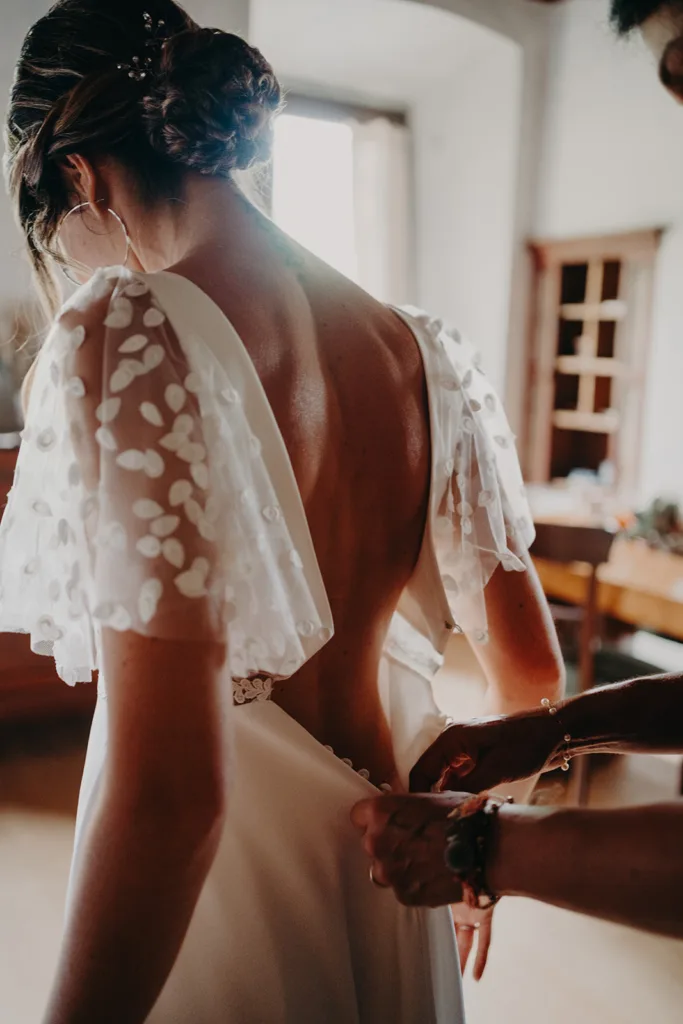 Une mariée vêtue d'une robe de mariée aux manches transparentes et fleuries a le dos de sa robe fermé par une autre personne dans une pièce chaleureusement éclairée, créant un moment intime semblable à une séance capturé sur la photo.