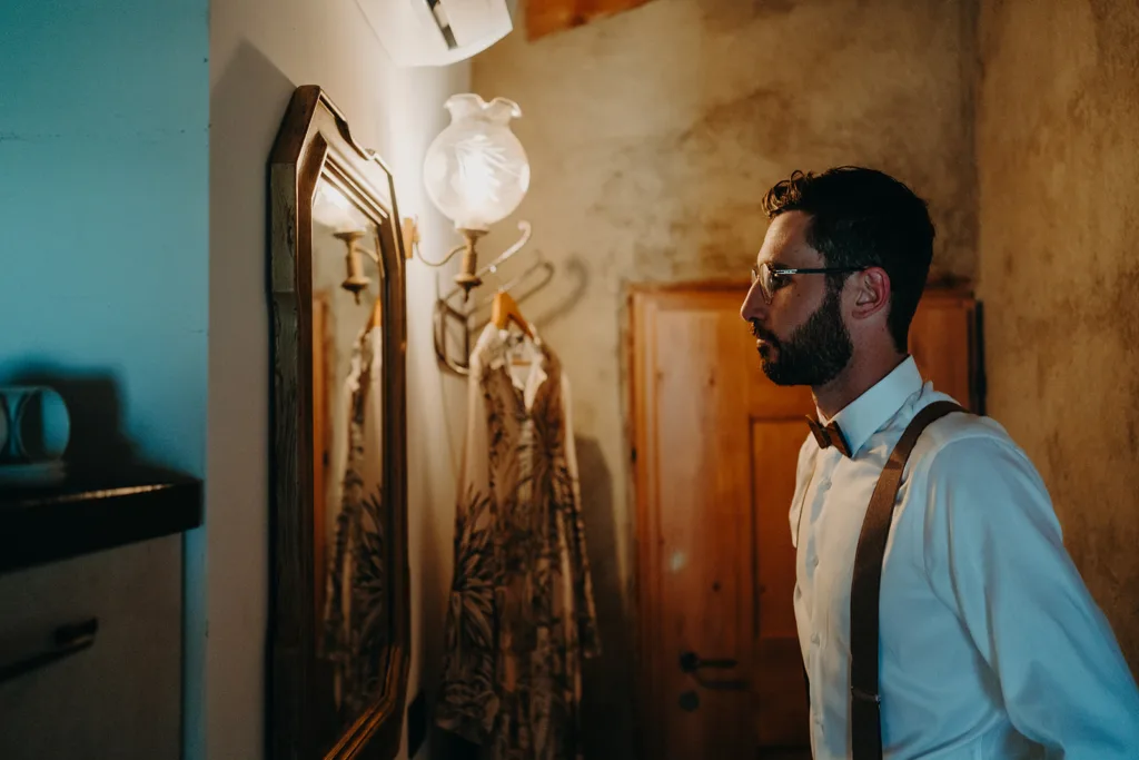 Un homme portant une barbe et des lunettes, vêtu d'une chemise blanche et de bretelles, se tient devant un miroir dans une pièce faiblement éclairée au décor rustique, rappelant une séance d'antan.