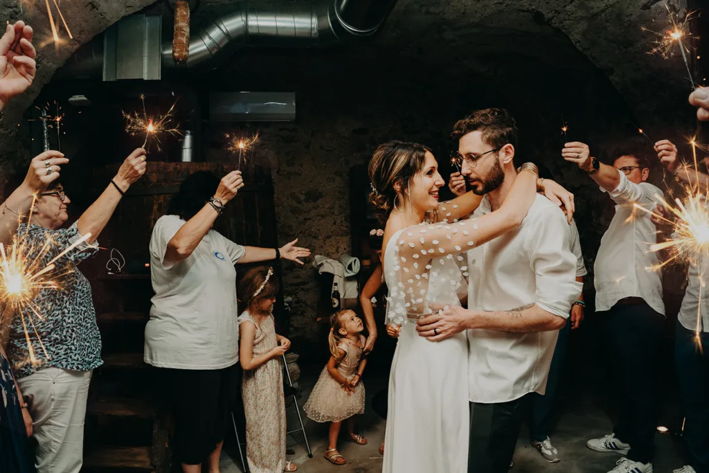 Un couple en tenue de mariage partage une danse entouré d'invités tenant des cierges magiques dans un espace faiblement éclairé, créant une scène enchanteresse qui ressemble presque à une photo de séance romantique.