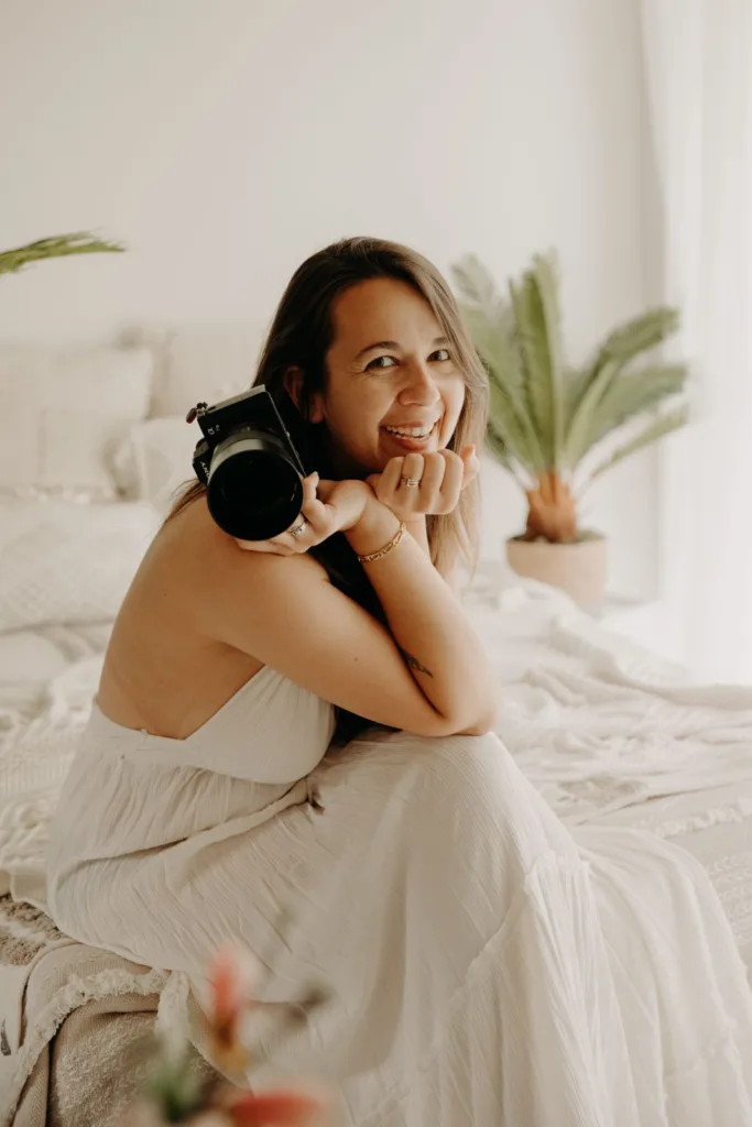 Photographe tenant un appareil photo et souriante, vêtue d'une tenue blanche sans manches avec des plantes d'intérieur en arrière-plan, capture l'essence décontractée d'un photographe de famille dans les Landes.